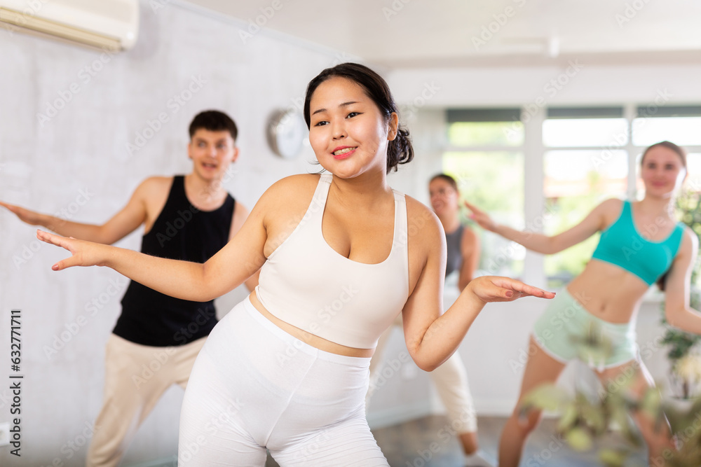 Asian female teen performs choreographic exercises and teaches energetic mobile social dance groove together with friends. Young girls and guy repeat movements, train in spacious studio