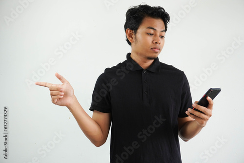 Young asian man holding a mobile phone isolated on white background serious and pointing aside, showing something at blank space, wearing black polo t shirt. photo