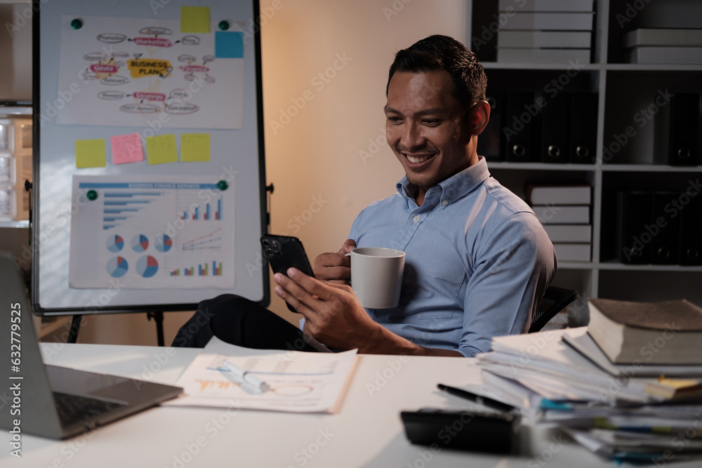 Fototapeta premium Smiling Asian businessman relaxing after work hard in the office, holding smartphone and coffee cup at office desk.