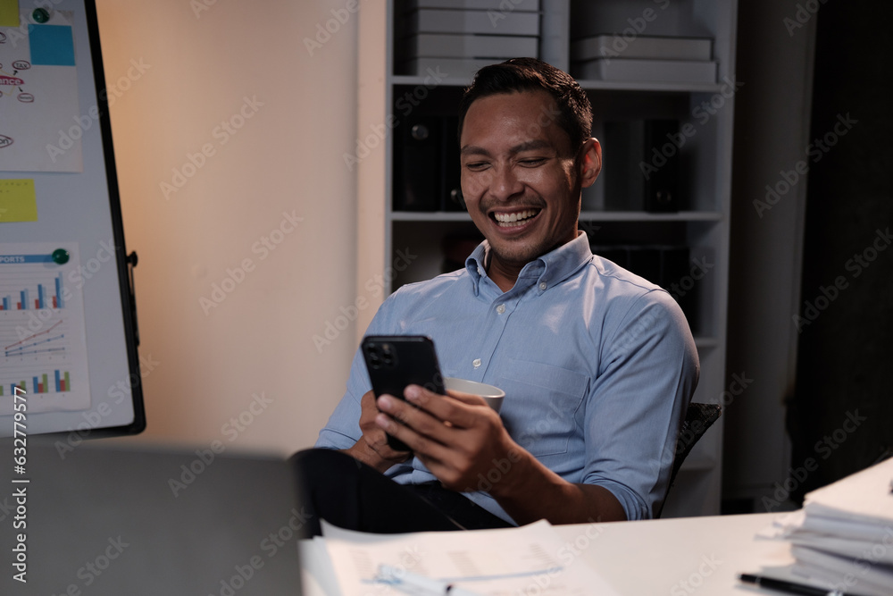 Fototapeta premium Smiling Asian businessman relaxing after work hard in the office, holding smartphone and coffee cup at office desk.