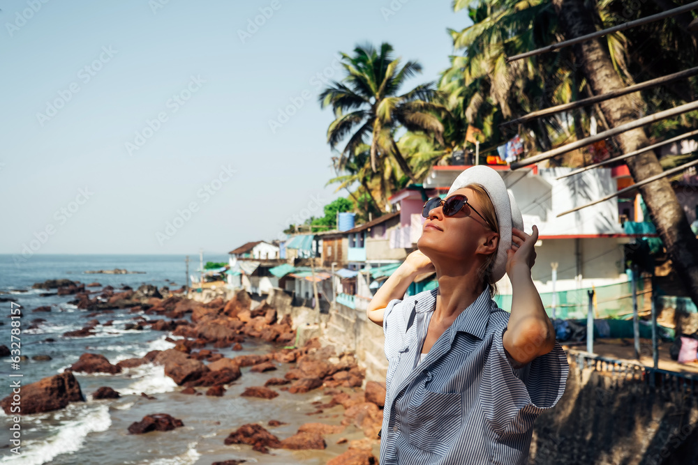 A beautiful tourist girl in dark glasses, a white hat and a blue shirt looks at the sea, palm trees and coastal houses. A concept for travel and tourism