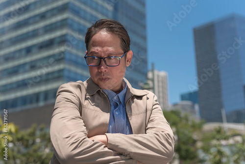A jaded middle aged asian man in his 40s looks down feeling dismayed and discouraged. Urban city background.