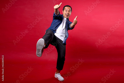 Asian young man in casual outfit showing thumb ups and raising leg up on blue background Asian young man in casual outfit showing thumb ups and raising leg up on blue background © Gatot