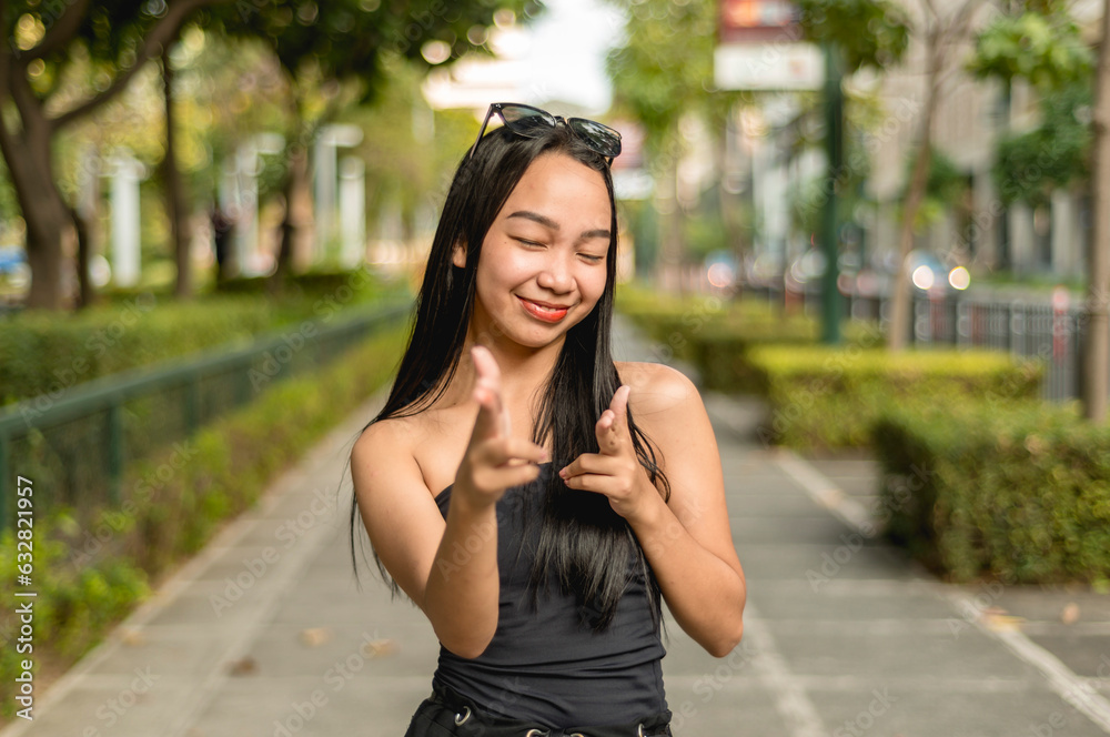 A pretty young lady standing in a park wearing a black strapless tank top and sunglasses, smiling and holding up two finger gun hand gesture. Trees,bushes and fence are in the background. A pretty young lady standing in a park wearing a black strapless tank top and sunglasses, smiling and holding up two finger gun hand gesture. Trees,bushes and fence are in the background.