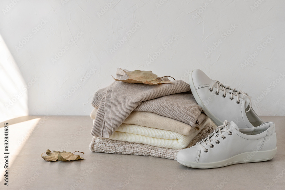 Fashion minimalist autumn clothes wardrobe in neutral beige colors. Beige, creamy, taupe wool knitted sweaters stack, white sneakers, brown fall leaves on table, on white empty wall background Fashion minimalist autumn clothes wardrobe in neutral beige colors. Beige, creamy, taupe wool knitted sweaters stack, white sneakers, brown fall leaves on table, on white empty wall background