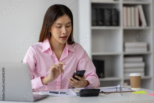 Asian businesswoman holding smartphone searching for information through website, online, surfing the internet with graph paper Work chart lying on the desk in the office