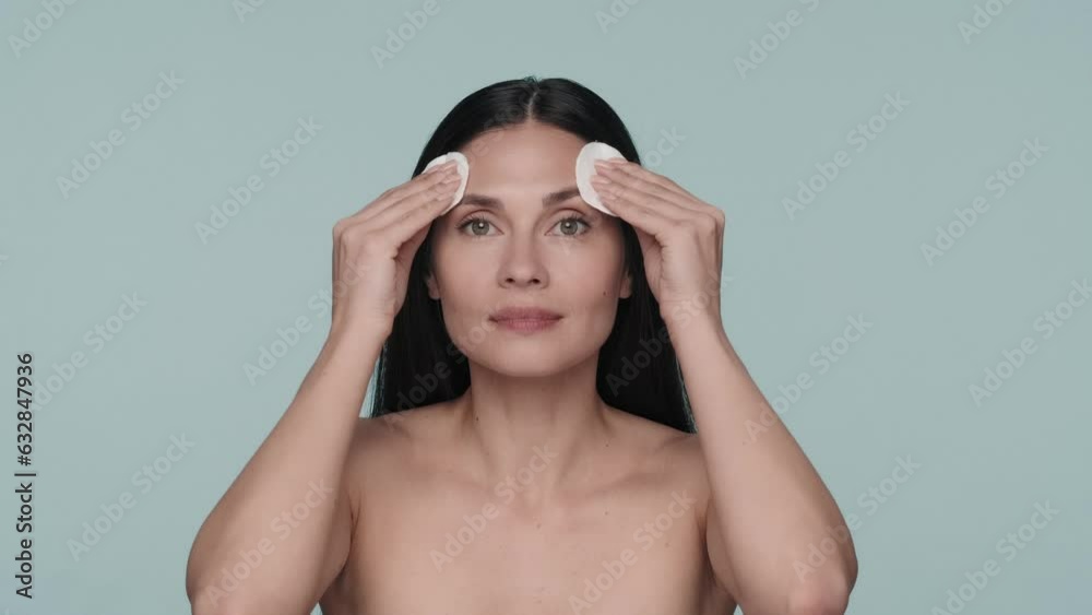 A woman takes care of her skin in the studio on a blue background close up. A seminude woman will wipe the skin of her face, neck and shoulders with the help of cotton pads. Slow motion. A woman takes care of her skin in the studio on a blue background close up. A seminude woman will wipe the skin of her face, neck and shoulders with the help of cotton pads. Slow motion.