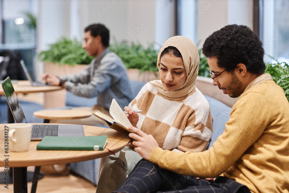 Portrait of modern Middle Eastern woman wearing head cover in office and looking at notes with male colleague Portrait of modern Middle Eastern woman wearing head cover in office and looking at notes with male colleague