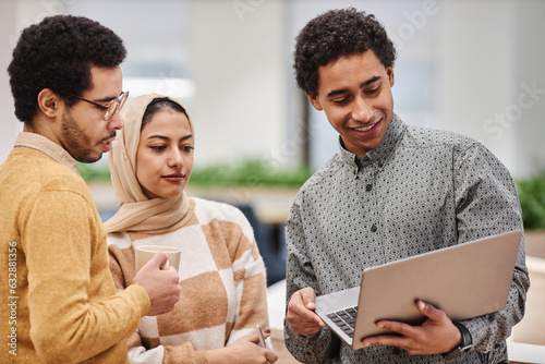 Group of modern Middle Eastern professionals holding laptop while discussing work project in office