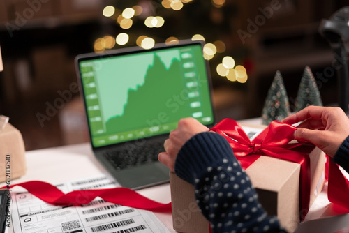 Female analyst at her desk works on a laptop showing statistics, graphs while working at Christmas. She Works on the Table packing package gift with red ribbon in creative office.