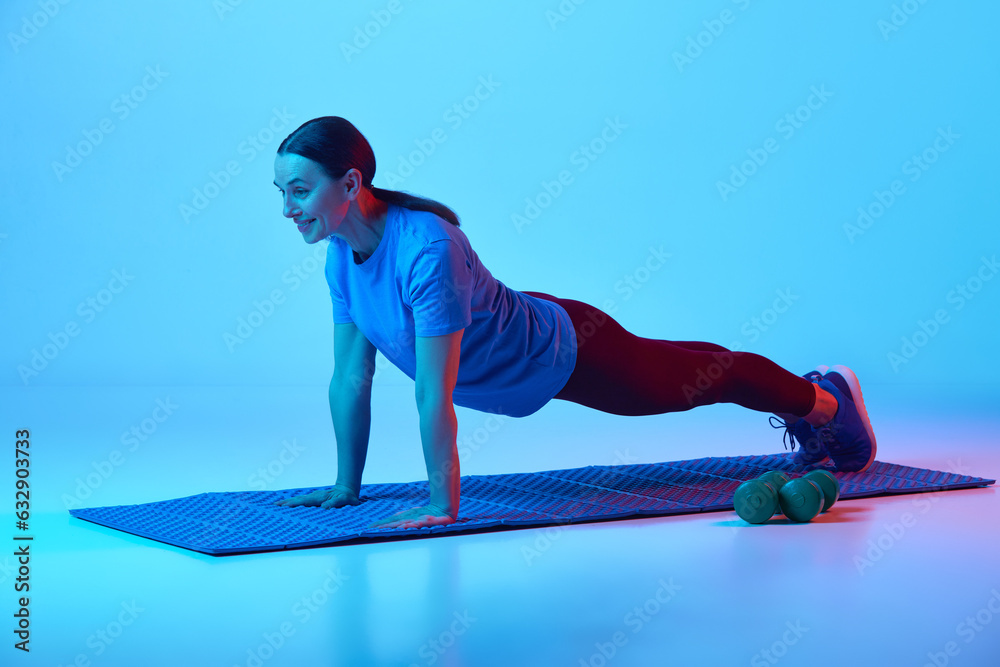 Beautiful mature woman training, standing in plank pose against blue studio background in neon light. Concept of sport, healthy lifestyle, fitness, body care, wellness, ad