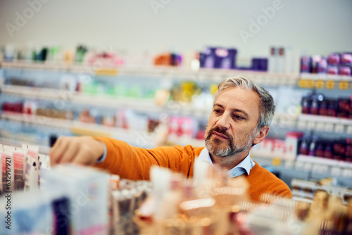 A senior man is standing by the shelf in a store and picking hand cream.
