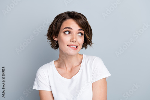 Photo of doubtful unsure woman dressed white t-shirt biting lip looking empty space isolated grey color background