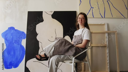Portrait of the artist against the background of his paintings. The woman painter smiles and looks into the camera. Artist's studio, canvases on the wall, abstract painting.