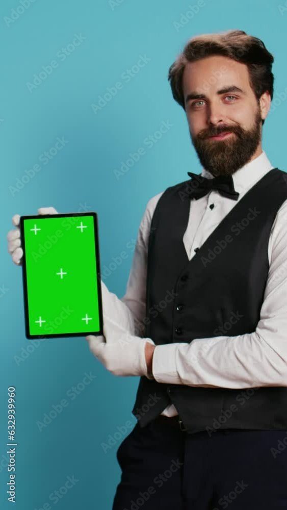 Vertical video Elegant bellboy holding tablet on camera and presenting blank greenscreen display, symbolizing hotel concierge services with professionalism. Porter in suit and tie pointing at isolated Vertical video Elegant bellboy holding tablet on camera and presenting blank greenscreen display, symbolizing hotel concierge services with professionalism. Porter in suit and tie pointing at isolated