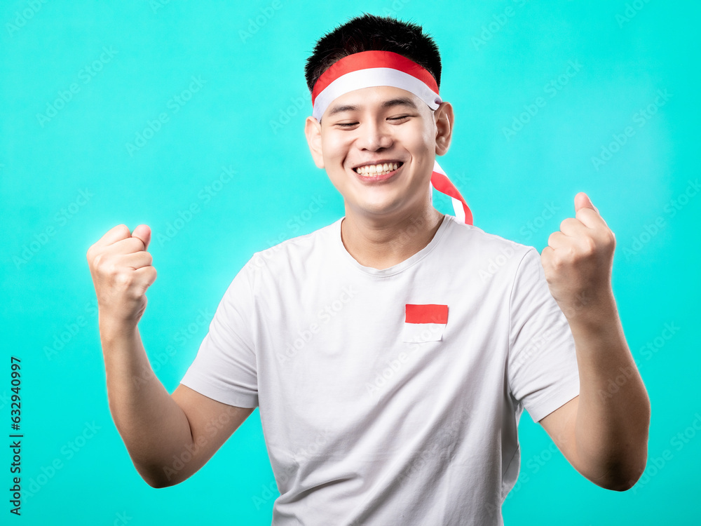 Naklejka premium A portrait of an Asian man celebrating Independence Day, wearing a white t-shirt and a red and white headband, showing a victory pose with his hands raised up. Isolated with a turquoise background.