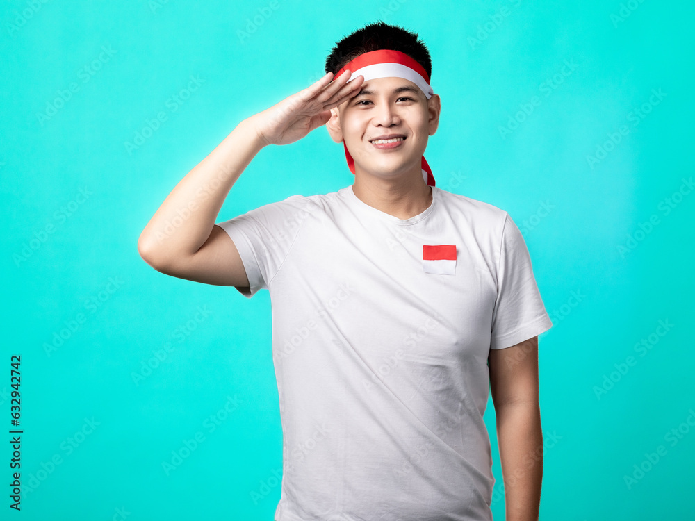 Fototapeta premium A portrait of an Indonesian (Asian) man celebrating Independence Day, wearing a white t-shirt and a red and white headband, in a salute pose. Isolated with a turquoise background.