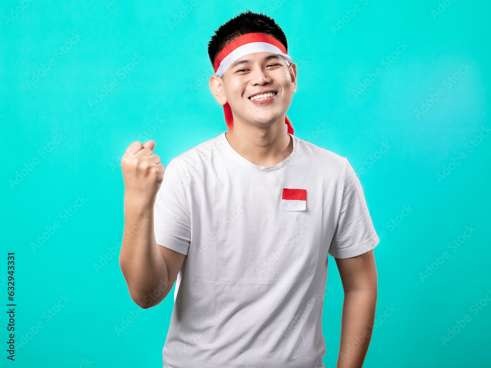 Fototapeta premium A portrait of an Asian man celebrating Independence Day, wearing a white t-shirt and a red and white headband, showing a victory pose with his hands raised up. Isolated with a turquoise background.