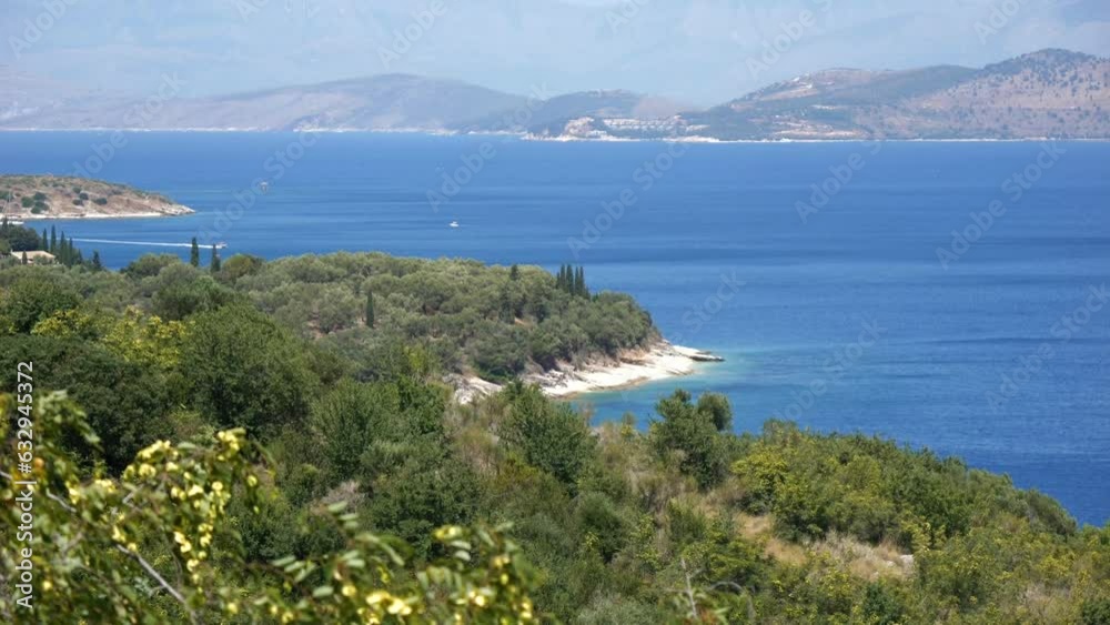 View of forested rocky cliffs on Corfu Island, with the blue sea and tall mountains in the background View of forested rocky cliffs on Corfu Island, with the blue sea and tall mountains in the background