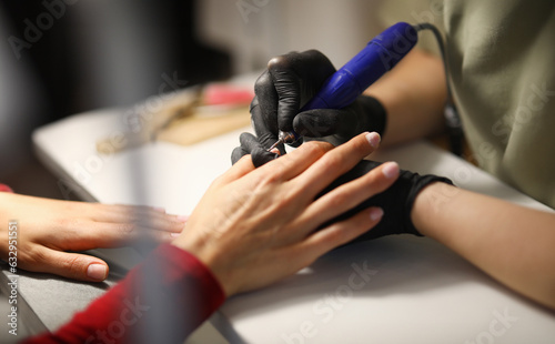 Professional in black gloves make mechanized manicure to visitor in studio. Woman processes her nails before applying varnish in workplace.