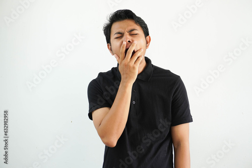 sleepy young asian man yawning by covering mouth with hand wearing black polo t shirt isolated on white background photo