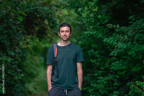 Happy Handsome beautiful man  30 years standing on road  forest green  shirt caucasian trees background outdoors nature park summer beard smiling happy casual backpack hands in pocket