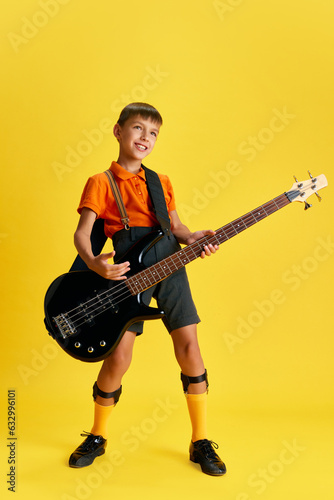 Boy, child in stylish clothes playing guitar against yellow studio background. Music and education. Concept of childhood, kids emotions, fashion, hobby, fashion and lifestyle, ad