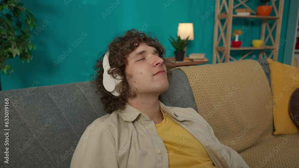 Young man in casual clothing and headphones sitting on the couch in the living room with closed eyes and listening to music
