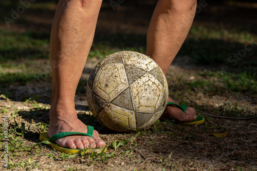 Mulher com os pés calçados com chinelo do Brasil verde e amarelo com uma bola de futebol entre os pés em campo de gramado gasto. photo