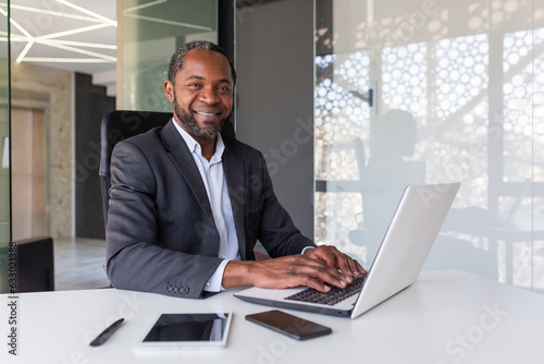 Portrait of successful happy african american boss, man smiling and looking at camera, businessman in business suit sitting at desk with laptop inside office.