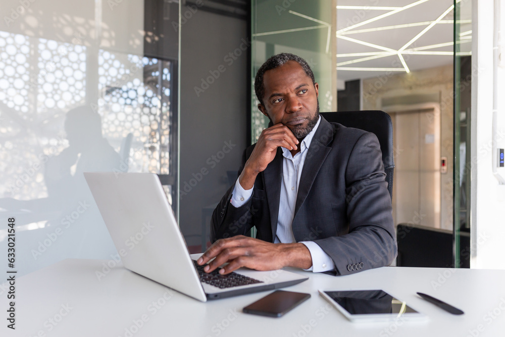 Thinking mature adult african american businessman at workplace, financier boss investor with laptop thinking about financial solutions new projects inside office.