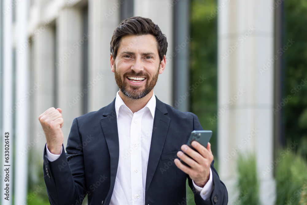 Portrait of a young businessman, lawyer holding and using the phone. Standing on the street and rejoicing in success, showing a victory gesture to the camera Portrait of a young businessman, lawyer holding and using the phone. Standing on the street and rejoicing in success, showing a victory gesture to the camera