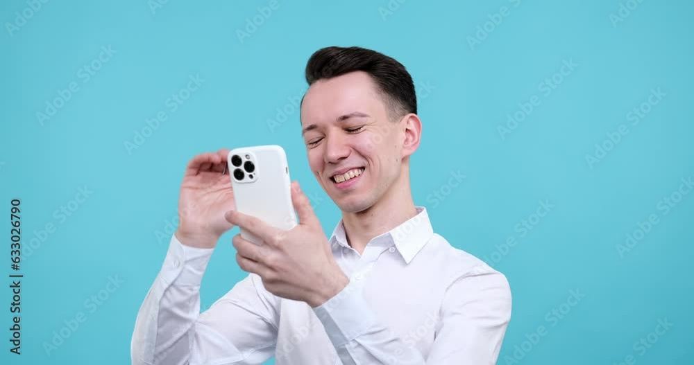 Caucasian man as he stands against a captivating blue background. He holds a smartphone in his hands, utilizing it for a video call. With a wide smile on his face, eyes light up with joy and surprise.