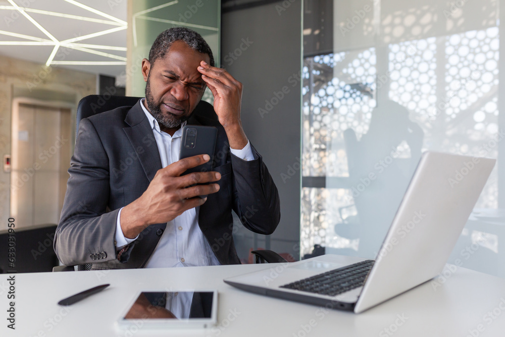 Upset and disappointed man inside office holding phone, businessman reading bad news at workplace inside office, african american senior boss unhappy with achievement results notification. Upset and disappointed man inside office holding phone, businessman reading bad news at workplace inside office, african american senior boss unhappy with achievement results notification.