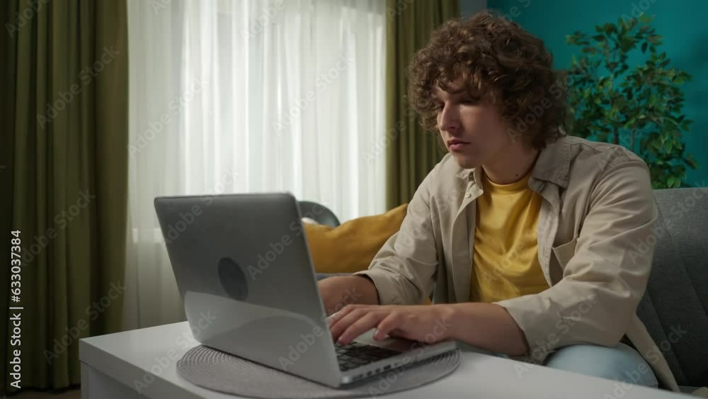 Young man in casual clothing, sitting on the couch in the living room and typing something on his laptop with serious face