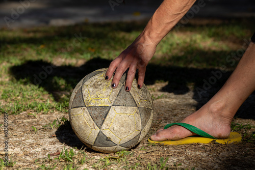 Detalhe da mão de uma mulher com as unhas pintadas ajeitando uma bola de futebol gasta e um de seus pés com chinelo verde e amarelo, se preparando para chutá-la. photo