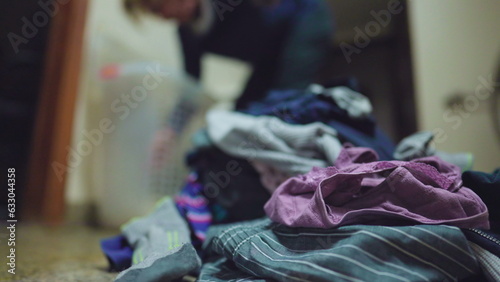 Domestic scene of person doing laundry. Close-up of clothes stacked together on floor with person blurred in background doing household routine