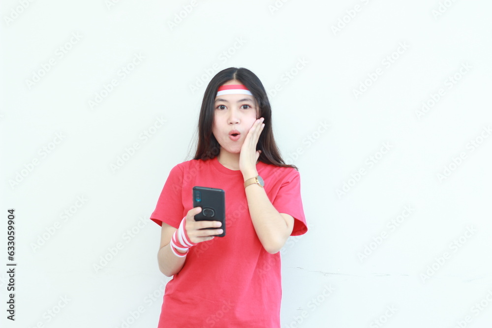 Naklejka premium portrait of beautiful asian woman wearing red outfit celebrating Indonesia independence day holding mobile phone with surprised expression