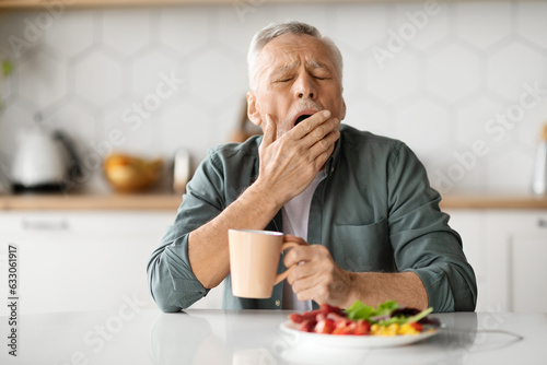 Excessive Daytime Sleepiness. Tired Senior Man Yawning At Table In Kitchen
