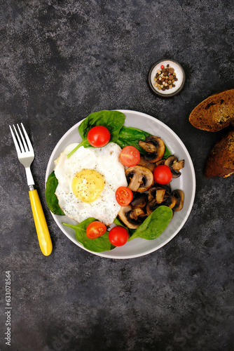 Fried egg, mushrooms, tomatoes and herbs with a yellow fork, on a dark background.