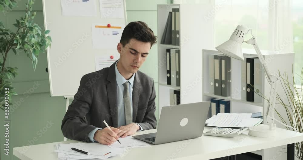 Stern Caucasian businessman is shown seated at his office desk, diligently writing on papers. With a focused and serious expression, he meticulously handles the documents. Stern Caucasian businessman is shown seated at his office desk, diligently writing on papers. With a focused and serious expression, he meticulously handles the documents.