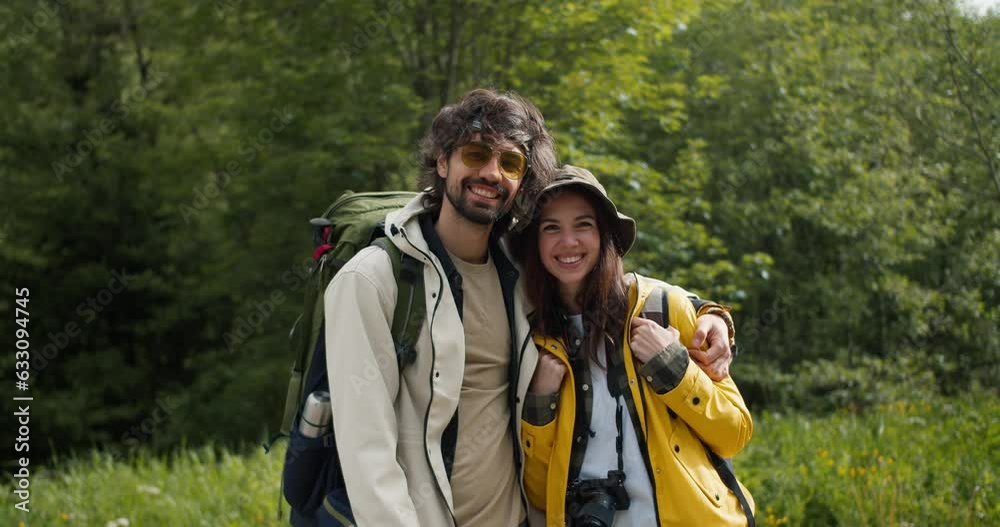 Portrait of a pair of brunette guy brunette girl in special clothes for hiking stand looking at the camera and posing against the backdrop of a green forest Portrait of a pair of brunette guy brunette girl in special clothes for hiking stand looking at the camera and posing against the backdrop of a green forest