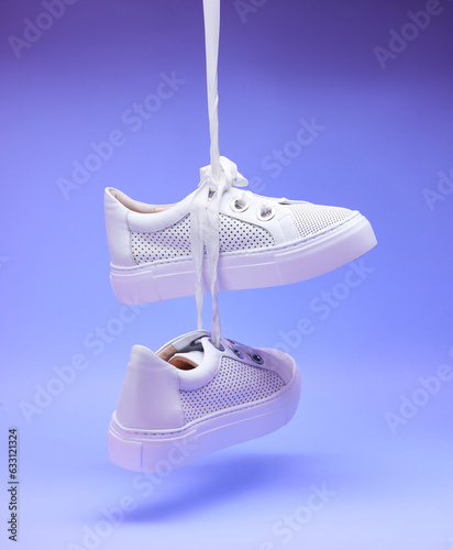 Casual white sneakers hanging on shoelaces in front of a gradient purple background. Perforated leather, thick sole, wide shoelaces, metal eyelets. Fashion blog or magazine concept. Neon light efect