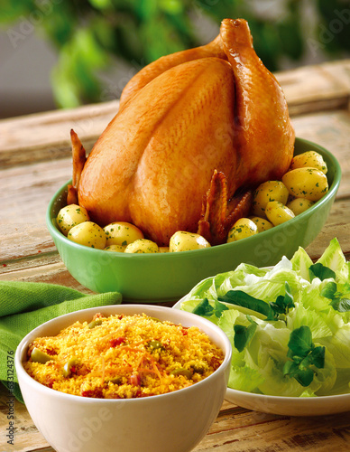 mesa de madeira rústica com frango assada com batatas acompanhado de receita brasileira de farofa e salada de folhas verdes