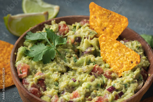 Wooden bowl of traditional Mexican guacamole with nachos on grey concrete background. Tortilla chips with guacamole sauce dip and ingredients: avocado, cilantro, onion, hot pepper and lime