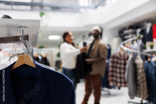 Clothes hanging on rack in store with bloggers taking selfie for brand promotion on blurred background. Casual apparel from new collection on hangers in fashion boutique close up selective focus