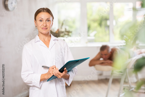 Smiling middle-aged masseuse holding documents standing in massage cabinet with her back to patient waiting for procedure