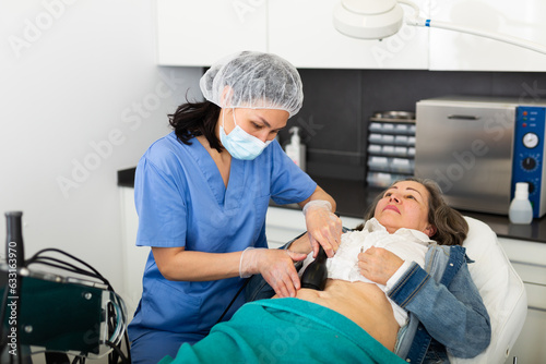 Asian woman professional cosmetologist performing abdominal skin rf lifting procedure on modern equipment for aged female patient in aesthetic medicine office