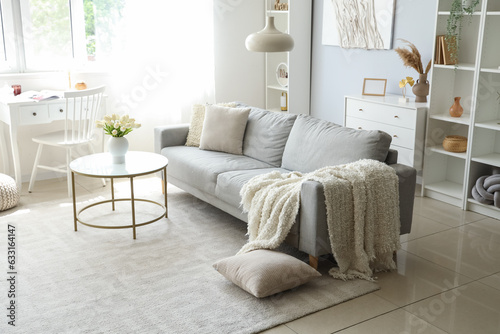 Cozy grey sofa with soft blanket and coffee table in interior of light living room