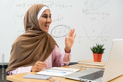 Young Eastern female teacher in hijab greeting students by waving hand during online lesson while sitting by desk in front of laptop. Smiling Islamic tutor in classroom. Education learning concept